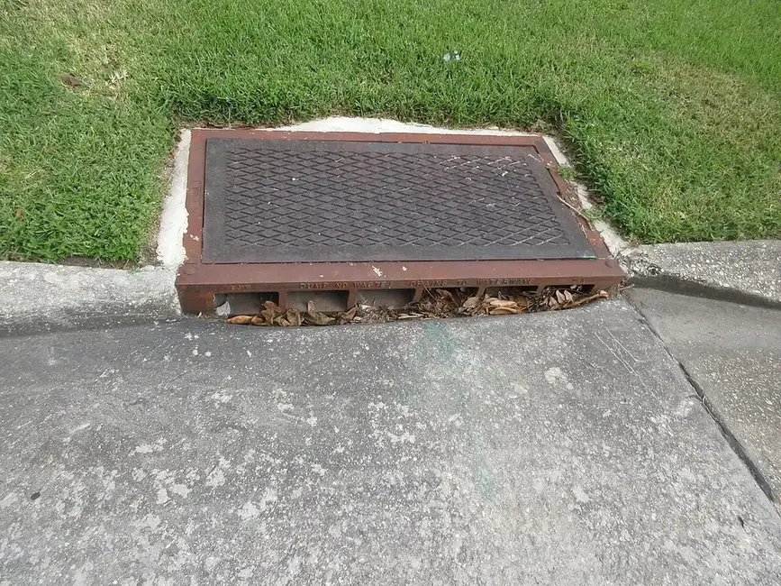 Cast iron catch basin grate set at the edge of a lawn where it meets a concrete apron