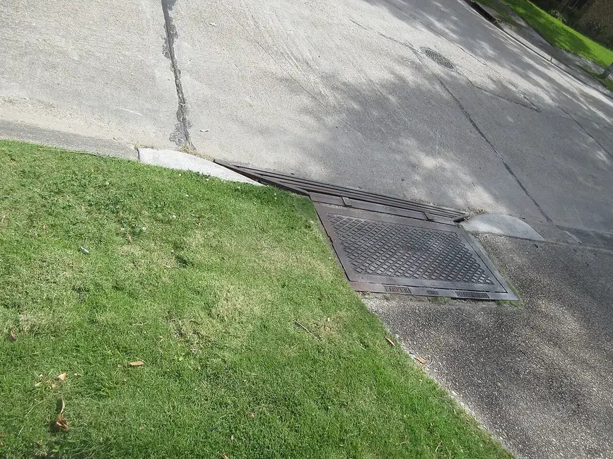 Cast iron catch basin grate and curb inlet where a sloped driveway meets the street beside a lawn