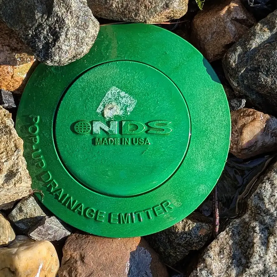 Green pop-up emitter lid at the end of a buried downspout drainage line, set among gravel