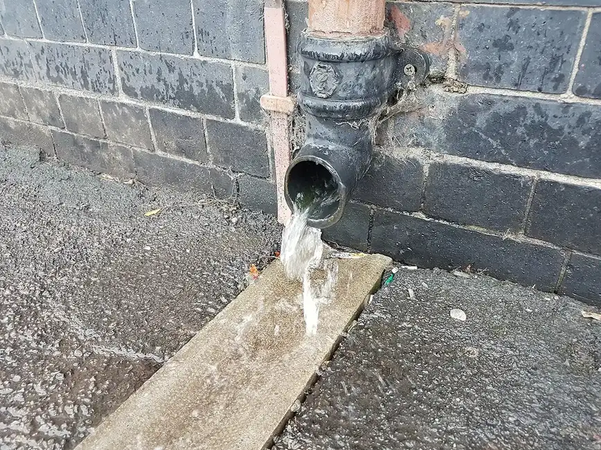 Downspout discharging water onto a wooden board on gravel right against a building wall