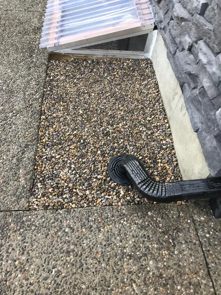 Black flexible downspout extension ending on pebbles beside a basement window well