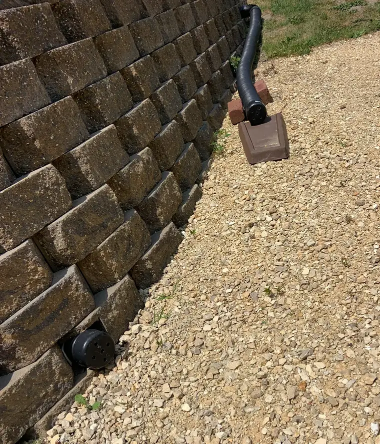 Black corrugated drain pipe on gravel at the foot of a stacked block retaining wall, with a round outlet cap at the wall's base