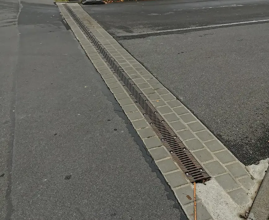 Cast iron trench drain grate running across a paved driveway entrance between asphalt sections